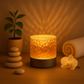 Lampe cylindrique texturée projetant une lumière dorée sur le mur, posée sur une surface en bois avec des galets empilés, une petite plante, une orchidée et une serviette roulée