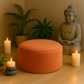 Coussin de méditation orange disposé dans un coin bien-être avec bougies, plantes et statue de Bouddha