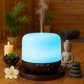 Diffuseur d’huiles essentielles au design bois et lumière bleue, posé dans une ambiance zen avec tapis de yoga, bougie, fleurs et statuette de Bouddha