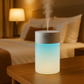 Diffuseur d’huiles essentielles au design cylindrique, émettant une lumière douce bleutée, posé sur une table de nuit dans une chambre cosy et relaxante.

