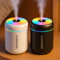 Deux humidificateurs d'air, l'un blanc et l'autre noir, avec éclairage LED arc-en-ciel diffusant de la vapeur, posés côte à côte sur une table en bois dans un environnement chaleureux.